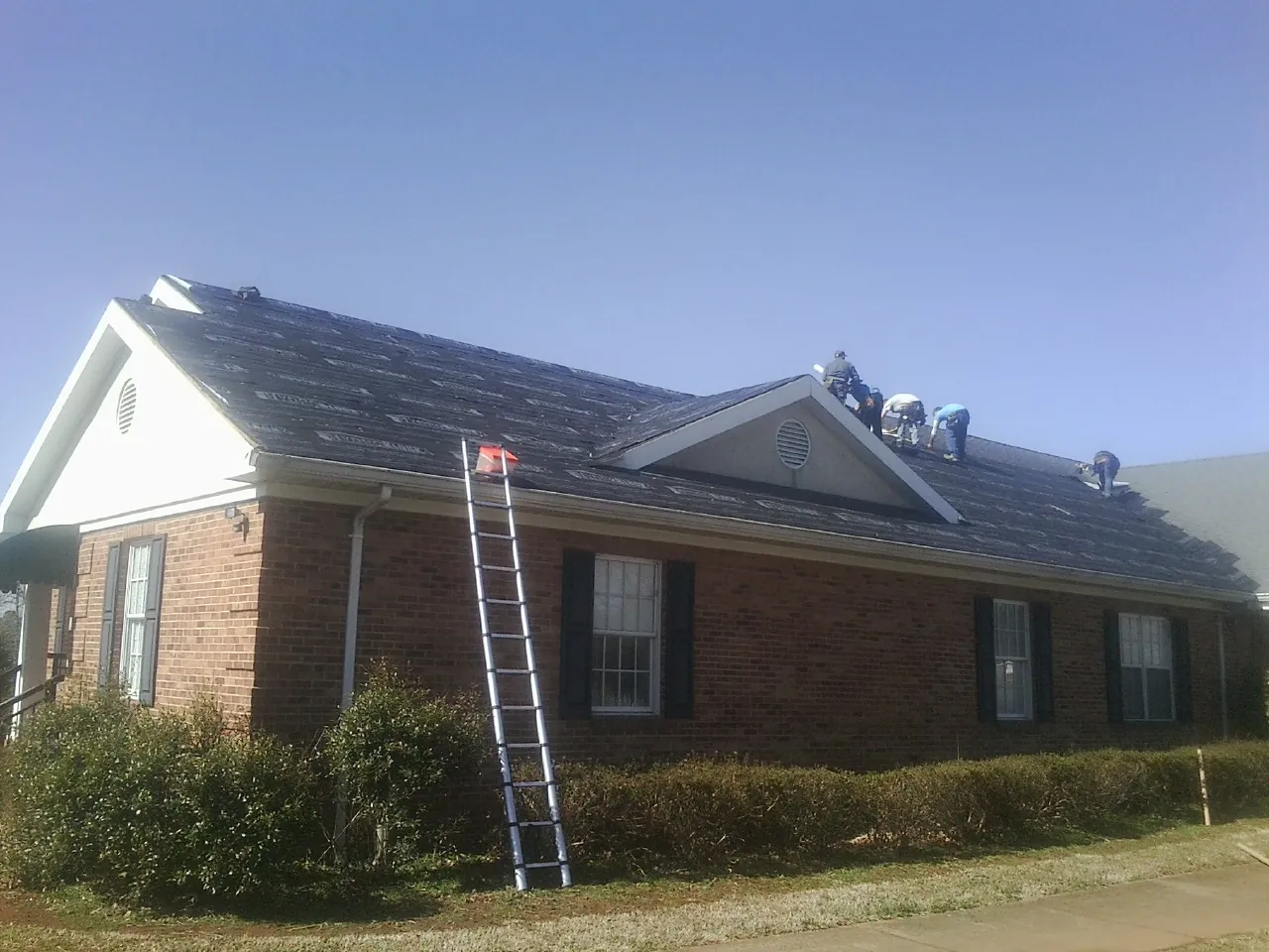 Crew tearing shingles off a brick ranch roof with a ladder set — Metal Roof Repair work by our crew in Lucernemines