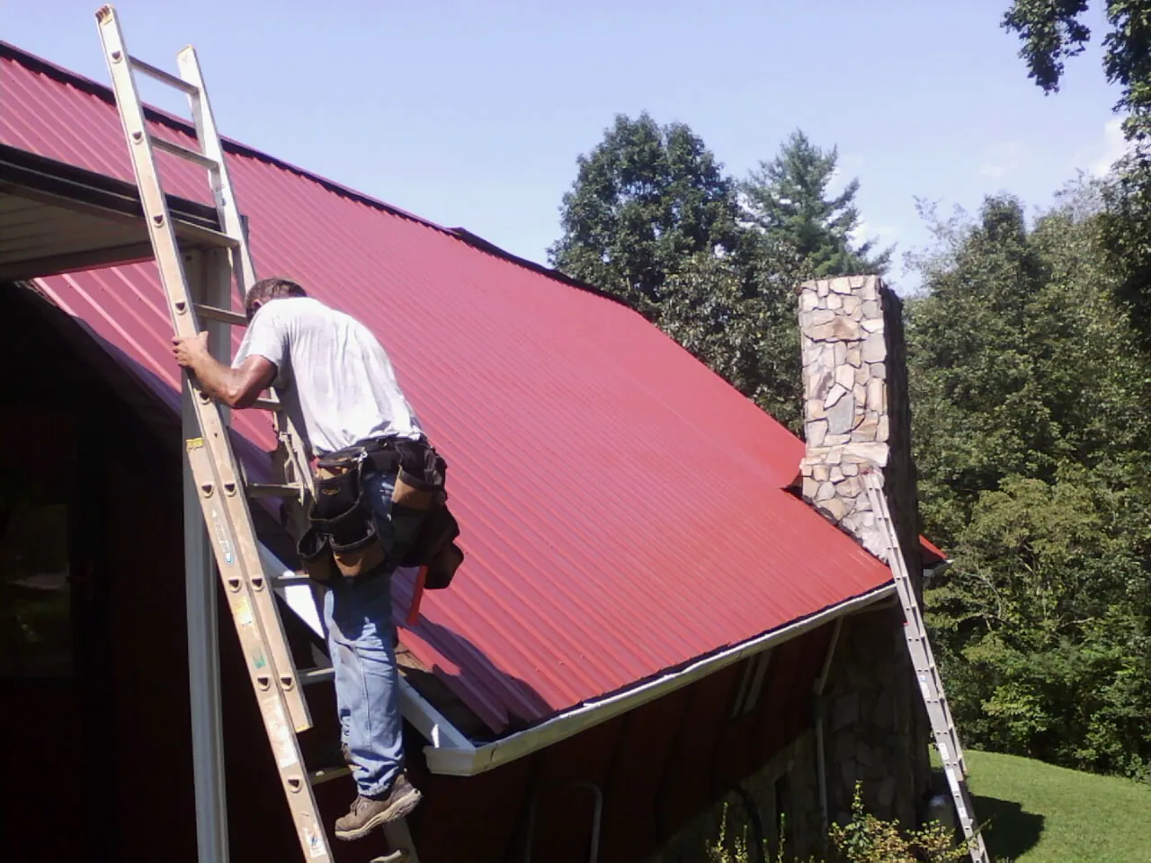 Roofer on a ladder beside a red metal roof and stone chimney — Soffit & Fascia Repair by Roofing Brothers in Lucernemines